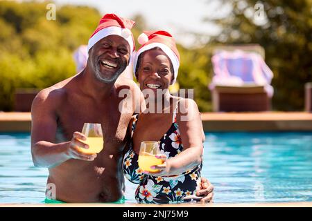 Ritratto di coppia anziano in vacanza di Natale in piscina indossando cappelli di Santa Foto Stock