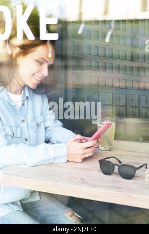 Donna seduta all'interno di un caffè al tavolo accanto alla finestra e utilizzando il suo telefono cellulare. Foto Stock