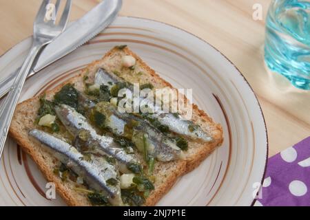 Una fetta di pane con acciughe condite con olio, aceto, aglio e prezzemolo su un piatto con posate vista dall'alto Foto Stock