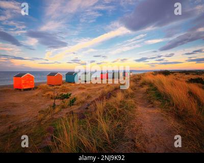 Un sentiero di sabbia conduce invitano attraverso le dune di Findhorn Beach, con le iconiche capanne sulla spiaggia che offrono un tocco di colore come le cascate del crepuscolo. Foto Stock