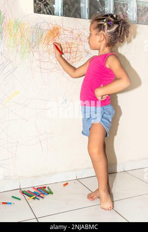 Piccola ragazza brasiliana che scarabocchio, dipinge e disegna su una parete di colore giallo chiaro Foto Stock