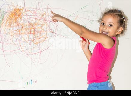 Piccola ragazza brasiliana che scarabocchio, dipinge e disegna su una parete di colore giallo chiaro Foto Stock