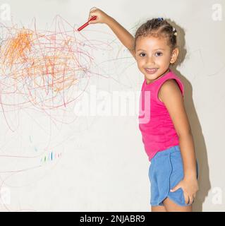 Piccola ragazza brasiliana che scarabocchio, dipinge e disegna su una parete di colore giallo chiaro Foto Stock