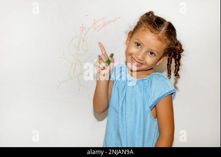 Piccola ragazza brasiliana che scarabocchio, dipinge e disegna su una parete di colore giallo chiaro Foto Stock