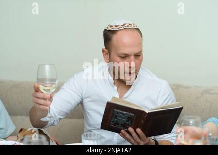 Giovane uomo beared con vino nel bicchiere che legge Haggadah. La famiglia ebraica festeggia la Pasqua al tavolo con cibo tradizionale. Cena con Pasqua in famiglia seder. Foto Stock