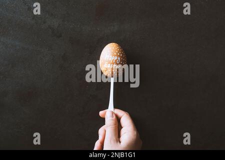 Mano che tiene un uovo scritto "Happy Easter" in un cucchiaio su fondo di metallo nero. Foto Stock