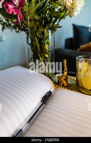 Piccolo blocco note A5 con linee si trova su un piccolo tavolino da caffè con matita nera accanto al vaso con fiori e piccola candela Foto Stock