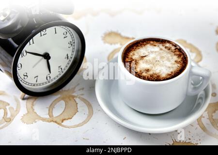 Una tazza di caffè, cappuccino e un orologio con un tavolo bianco con macchie di caffè Foto Stock