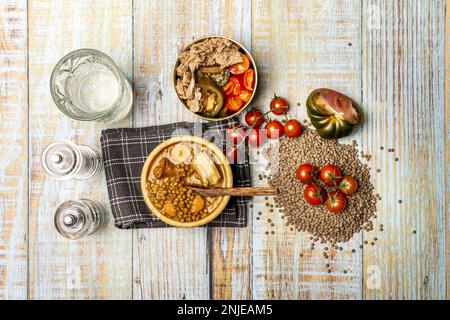 Vita morta con tradizionale stufato di lenticchie in una ciotola di legno e insalata di tonno e pomodoro in una ciotola dorata su un tavolo di legno chiaro con vasetti di spezie, len secco Foto Stock