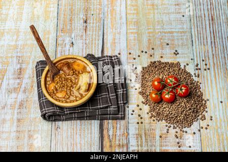 Vita morta con tradizionale lenticchie stufato in una ciotola di legno su un tavolo di legno chiaro con lenticchie secche e corde di pomodori ciliegini Foto Stock