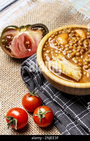 Ancora vita con tradizionale lenticchie stufato in una ciotola di legno su un tavolo di legno chiaro con una stringa di pomodori ciliegini e un pomodoro di marmellata dolce su assort Foto Stock