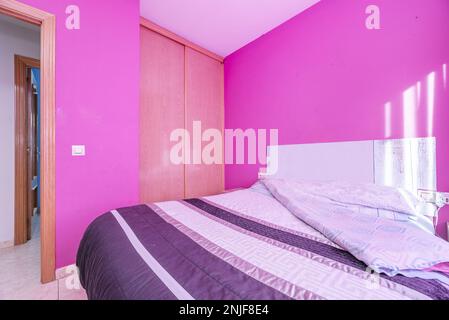 Camera con letto matrimoniale con copriletto viola, pareti rosa e armadio a muro con porte scorrevoli in rovere Foto Stock