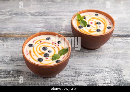 Yogurt con mirtillo e caramello in recipiente di argilla su fondo di legno grigio. vista laterale, primo piano. Foto Stock