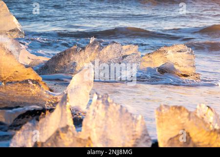 Ghiaccio del ghiacciaio puro che splende al tramonto. Sabbia vulcanica nera sulla costa dell'oceano. Onde oceaniche pure schiantano iceberg dorati in Islanda. Diamond Beach - famosa Foto Stock
