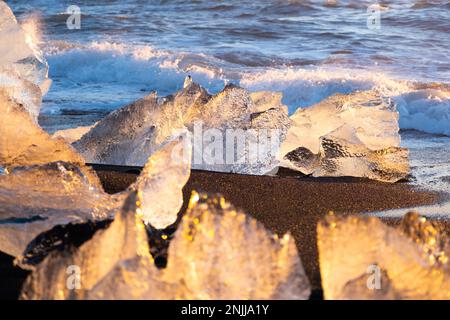 Ghiaccio del ghiacciaio puro che splende al tramonto. Sabbia vulcanica nera sulla costa dell'oceano. Onde oceaniche pure schiantano iceberg dorati in Islanda. Diamond Beach - famosa Foto Stock