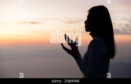 Silhouette of Woman che prega per ringraziare dio che prega con le sue mani insieme per pensare ad un Dio amorevole, noi lodiamo Dio con luce bagliante nella natura verde Foto Stock