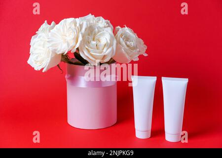 Profumo di rosa bianco naturale. Tubo di shampoo per capelli bianco vuoto con flacone di siero e rose biologiche fresche su sfondo rosso. Concetto di bellezza di cura dei capelli di erbe Foto Stock