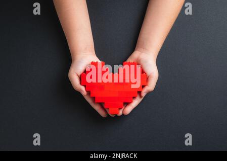 Mani di bambina che tiene il cuore fatto di mattoni di plastica su sfondo grigio scuro Foto Stock