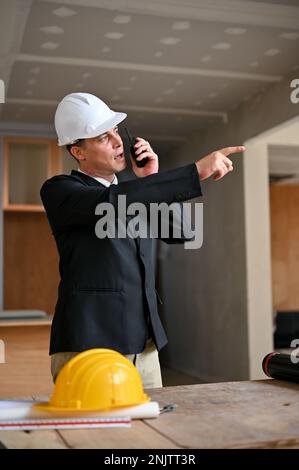 Ritratto, determinato e professionale uomo d'affari caucasico in un hardHat bianco e tuta d'affari formale utilizzando un walkie-talkie per comandare i suoi lavoratori a. Foto Stock