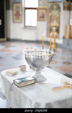 Fonte battesimale con acqua di benedizione, candele di cera e croce sono sul tavolo coperto da tovaglia bianca vicino alla Sacra Bibbia su sfondo icone. Newb Foto Stock