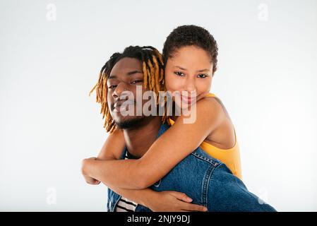 Ragazzo afro-americano positivo con capelli tinti in greggi in giacca denim dando piggyback cavalcare alla ragazza sorridente in top giallo mentre looki Foto Stock