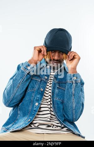 Giovane afro-americano positivo con baffi e lucchetti in giacca denim seduta su sedia e cappello a trazione sulla fronte in studio Foto Stock