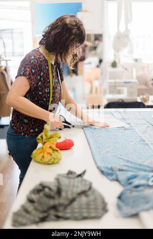 Vista laterale di una donna designer focalizzata in abbigliamento casual taglio pezzo di tessuto con forbici mentre si lavora sul modello creativo in officina Foto Stock