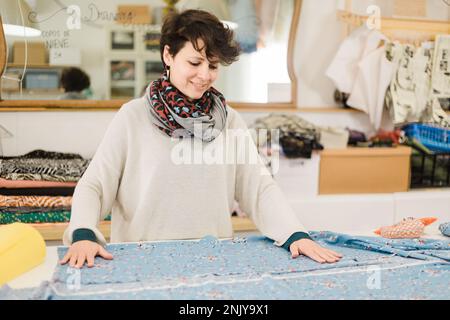 Artigiana sorridente focalizzata con metro a nastro sul collo per preparare il tessuto per creare un motivo in atelier Foto Stock