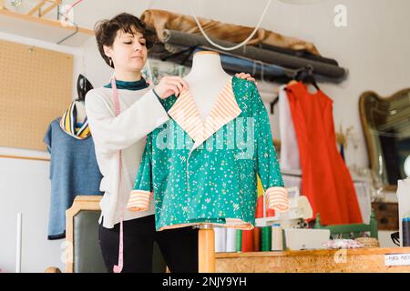 Sartoriale femminile concentrato in abiti casual con nastro di misurazione sul collo in piedi vicino al manichino mentre si prepara l'abbigliamento per il lavoro in studio creativo Foto Stock