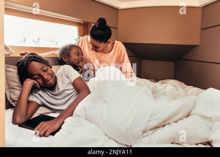 Positiva madre afro-americana sorridente e seduta a letto mentre si riposa e gioca con i bambini durante il fine settimana Foto Stock