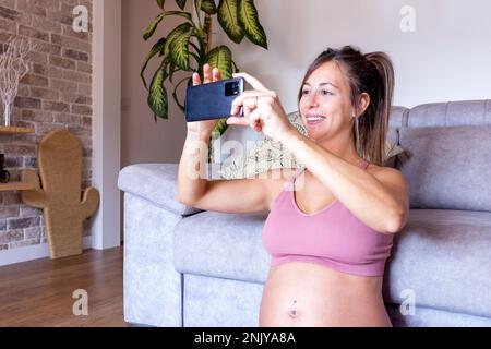 Felice giovane donna incinta in top rosa sorridente e scattando foto sullo smartphone mentre si siede sul pavimento vicino al divano a casa Foto Stock