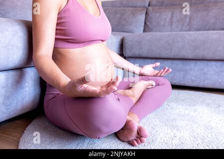 Corto irriconoscibile giovane donna incinta rilassata in rosa sportswear seduto sul pavimento vicino al divano e facendo esercizi di respirazione con palmi rivolti verso l'alto Foto Stock