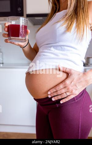 Vista laterale di una donna incinta tagliata e irriconoscibile nella parte superiore bianca che tocca il ventre e beve succo rosso fresco mentre si è in piedi al bancone della cucina Foto Stock