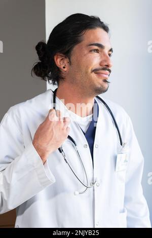 Giovane medico etnico sorridente in uniforme bianca con stetoscopio sul collo in piedi in clinica leggera Foto Stock