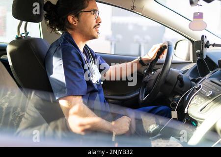 Vista laterale di un medico etnico maschile focalizzato in uniforme medica blu con stetoscopio e occhiali che guidano l'auto in città Foto Stock