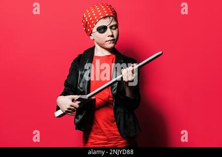 Ragazzo carino in vestito pirata giocando con la spada e guardando giù contro sfondo rosso brillante Foto Stock