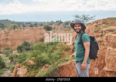Vista laterale di sorridente turista maschile in cappello con zaino ammirando il pittoresco paesaggio di valle sabbiosa del deserto in Colombia vicino al verde cactus guardare Foto Stock