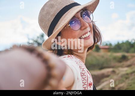 Donna allegra in cappello di paglia e occhiali sorridenti mentre tiene mano di raccolto anonimo partner al deserto Tatacoa in Colombia Foto Stock