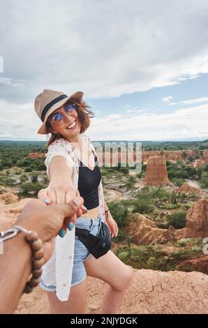 Donna allegra in cappello di paglia e occhiali sorridenti guardando la macchina fotografica mentre tiene mano di raccolto anonimo partner a Tatacoa deserto in Colombia Foto Stock