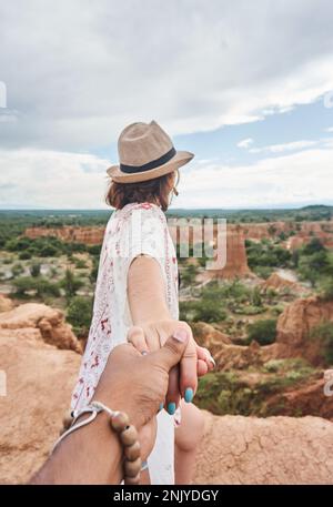 Vista posteriore della donna irriconoscibile in cappello di paglia e occhiali sorridenti guardando la macchina fotografica mentre tiene mano di raccolto anonimo partner a Tatacoa Desert Foto Stock