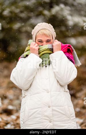 Donna adulta felice in abiti caldi e cappello che si nasconde dietro la sciarpa mentre si passa il tempo nel parco durante le nevicate Foto Stock