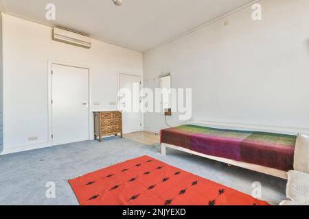 Design interno di spaziosa camera da letto con moquette sul pavimento e tappeto rosso vicino al letto singolo con specchio appeso su parete bianca Foto Stock