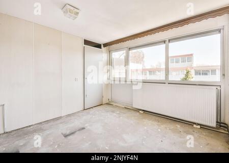 Design interno di camera spaziosa e luminosa con grandi finestre e radiatori con nuove carte da parati beige sulle vecchie pareti e pavimento in cemento Foto Stock