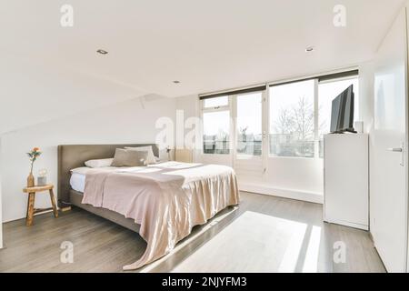 Moderno design interno di camera da letto chiara con letto king size vicino alla poltrona con tavolo e grande vaso pianta con radiatore vicino alla finestra in soffitta della casa Foto Stock