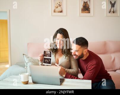 Felice marito bearded abbracciare sorridente moglie incinta wile seduto sul divano rosa vicino al computer portatile e mostrando l'immagine di ultrasuoni durante la videochiamata Foto Stock