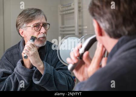 Uomini anziani concentrati in occhiali che guardano lo specchio e la barba rasata con rasoio elettrico in bagno Foto Stock