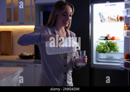 Giovane donna che versa il latte dalla bottiglia di gallone nel vetro vicino al frigorifero in cucina di notte Foto Stock