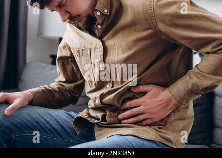 Rovesciare il maschio schiacciando la pancia con la mano a causa del dolore addominale. Un giovane infelice si siede sul divano grigio a casa, sentendo improvvisamente forte mal di stomaco, gas Foto Stock