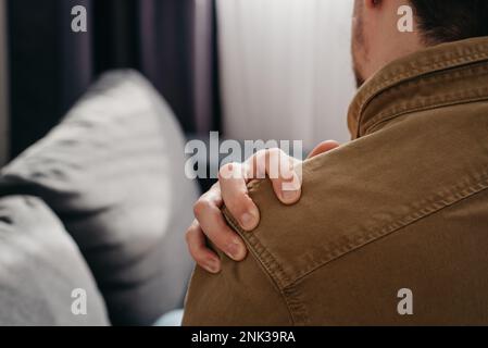 Dolore alla spalla. Dolore alla parte superiore del braccio, persone con problemi di corpo-muscoli, salute e concetto di medicina. Primo piano vista posteriore del giovane seduto sul divano A. Foto Stock