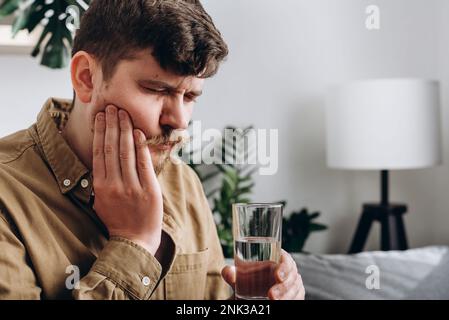 Primo piano di un giovane triste sentire tremendo mal di denti dopo aver bevuto acqua fredda. Maschio 30s seduto sul divano a casa toccare guancia, sentirsi male e sofferenza per il fro Foto Stock